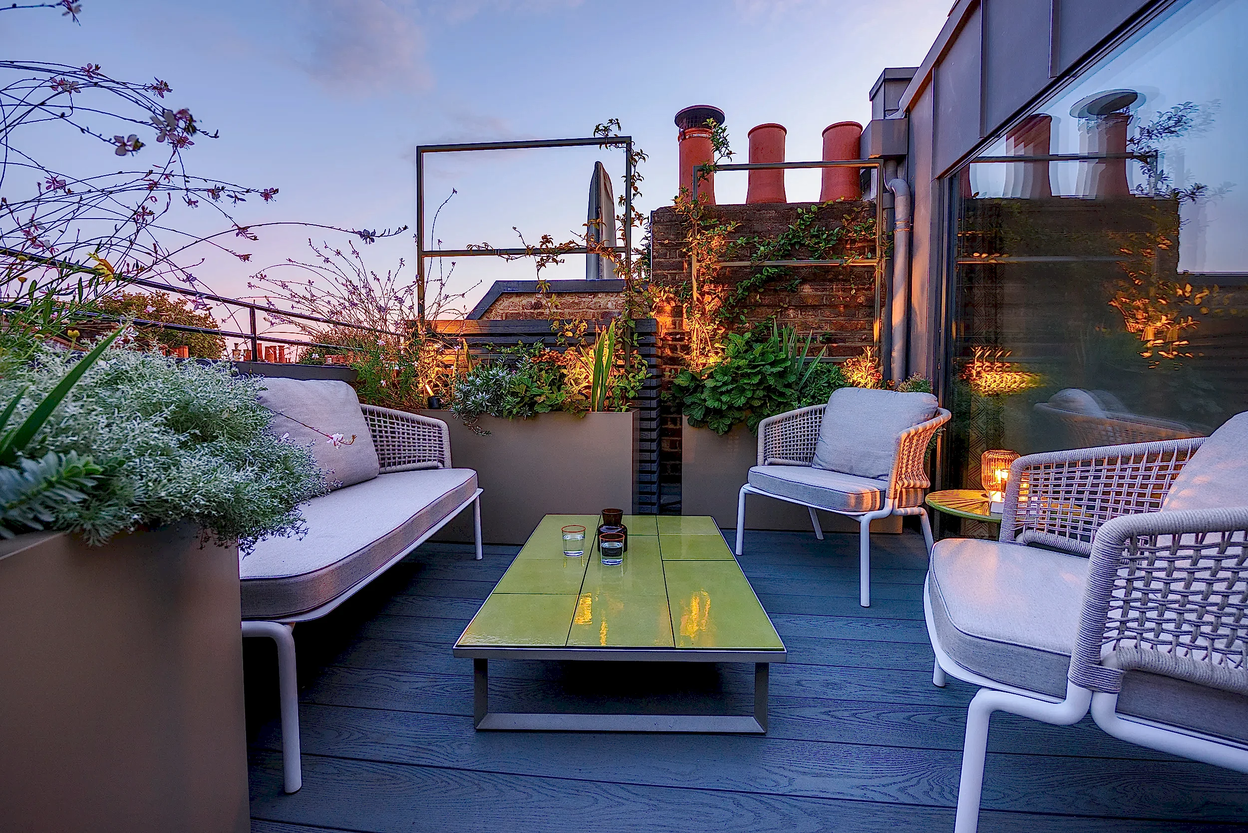 A London roof terrace at evening time illuminated by lights hidden in planters. Cream coloured seating is shouldered by overflowing planters. Center of the image is a green tiled coffee table.